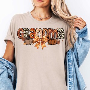 May include: A beige t-shirt with the word "Grandma" in colorful patterned letters. The letters are decorated with fall-themed elements like pumpkins, leaves, and a bow.