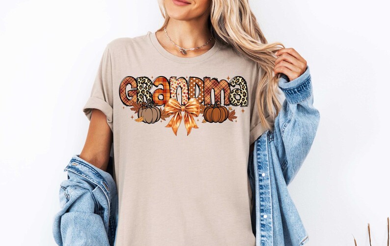 May include: A beige t-shirt with the word "Grandma" in colorful patterned letters. The letters are decorated with fall-themed elements like pumpkins, leaves, and a bow.