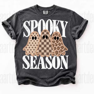 May include: A charcoal grey short-sleeve t-shirt featuring the white distressed text SPOOKY SEASON. The graphic includes three ghosts with black eyes, each with a different pattern: one with brown polka dots, one with a brown checkerboard, and one with leopard print.