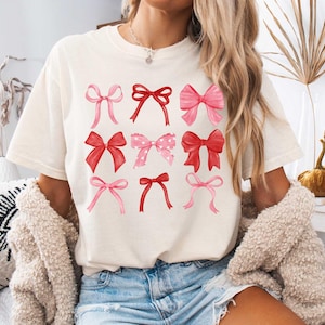 May include: A white t-shirt with a graphic of pink and red bows. The bows are arranged in a grid pattern on the shirt.