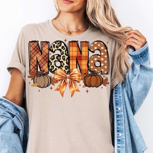 May include: A beige t-shirt with a graphic design of the word "NANA" in a patchwork style with fall colors, pumpkins, and a bow.