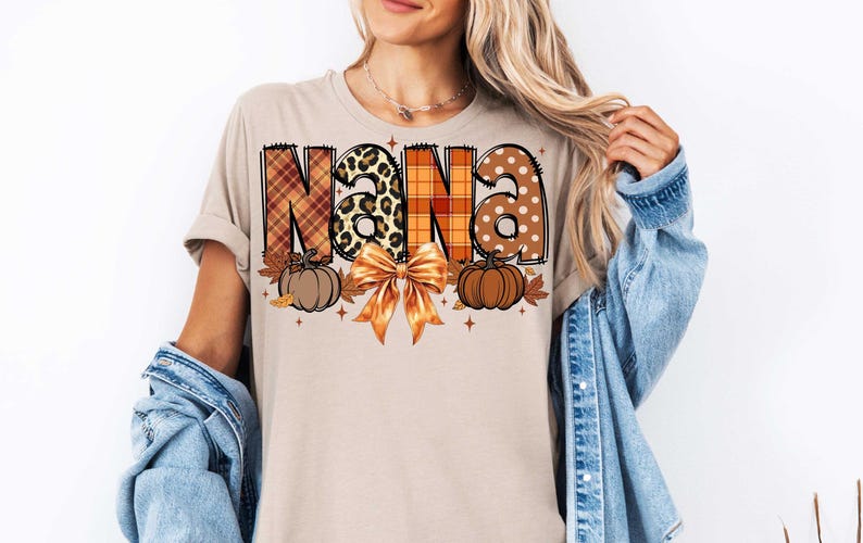 May include: A beige t-shirt with a graphic design of the word "NANA" in a patchwork style with fall colors, pumpkins, and a bow.