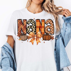 May include: White t-shirt with the word "NANA" in orange, brown, and black plaid, leopard print, and white polka dot patterns. The letters are decorated with fall leaves and pumpkins.