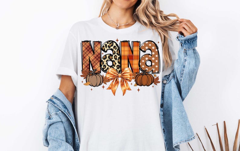 May include: White t-shirt with the word "NANA" in orange, brown, and black plaid, leopard print, and white polka dot patterns. The letters are decorated with fall leaves and pumpkins.