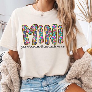 May include: A white t-shirt with the word "MINI" in colorful floral letters. The names "Jasmine", "Alex", and "Bruno" are written below the word "MINI" in black.