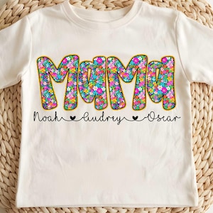 May include: White t-shirt with a floral design that says "Mama" in large letters. Below the word "Mama" are the names "Noah", "Audrey", and "Oscar" with hearts between each name.