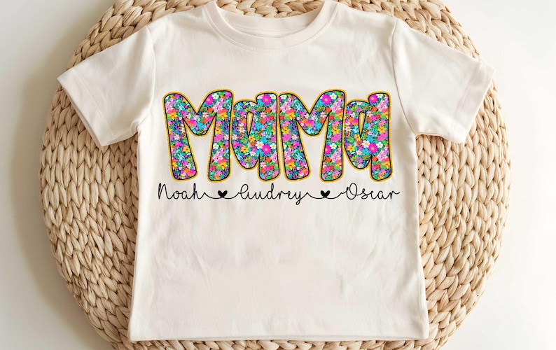 May include: White t-shirt with a floral design that says "Mama" in large letters. Below the word "Mama" are the names "Noah", "Audrey", and "Oscar" with hearts between each name.