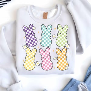 May include: White sweatshirt with a checkered pattern Easter bunny design. The bunnies are in various pastel colors with white flowers and sparkles.