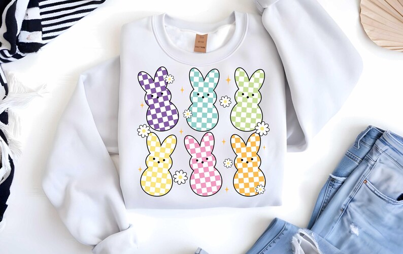 May include: White sweatshirt with a checkered pattern Easter bunny design. The bunnies are in various pastel colors with white flowers and sparkles.