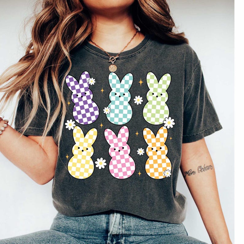 May include: A dark gray t-shirt with a colorful checkered bunny rabbit design. The bunnies are in a variety of colors, including purple, blue, green, yellow, and pink. The shirt has a crew neck and short sleeves.