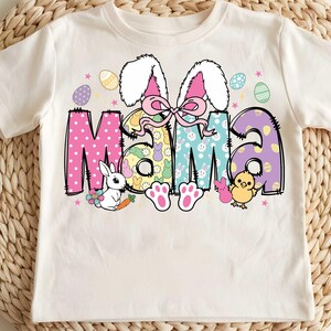 Pu&ograve; includere: Una maglietta bianca con un design colorato a tema pasquale che presenta la parola "Mama" circondata da coniglietti, uova e altre decorazioni pasquali.