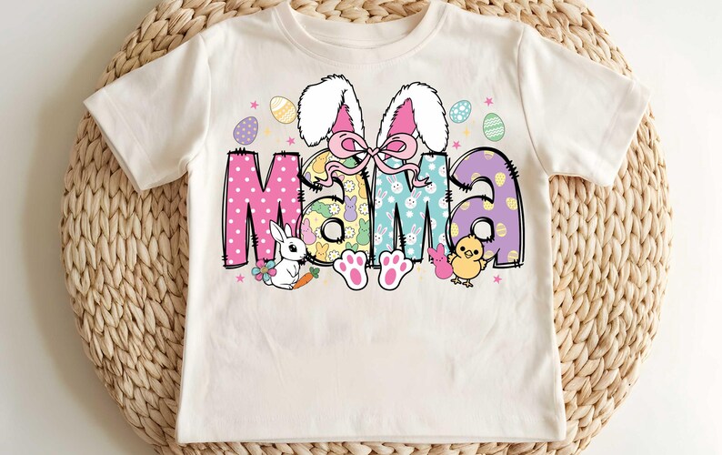 Pu&ograve; includere: Una maglietta bianca con un design colorato a tema pasquale che presenta la parola "Mama" circondata da coniglietti, uova e altre decorazioni pasquali.