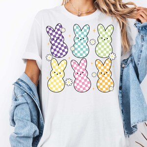 May include: White t-shirt with a graphic of six pastel colored Easter bunnies with checkered patterns. Each bunny has a small flower behind it.