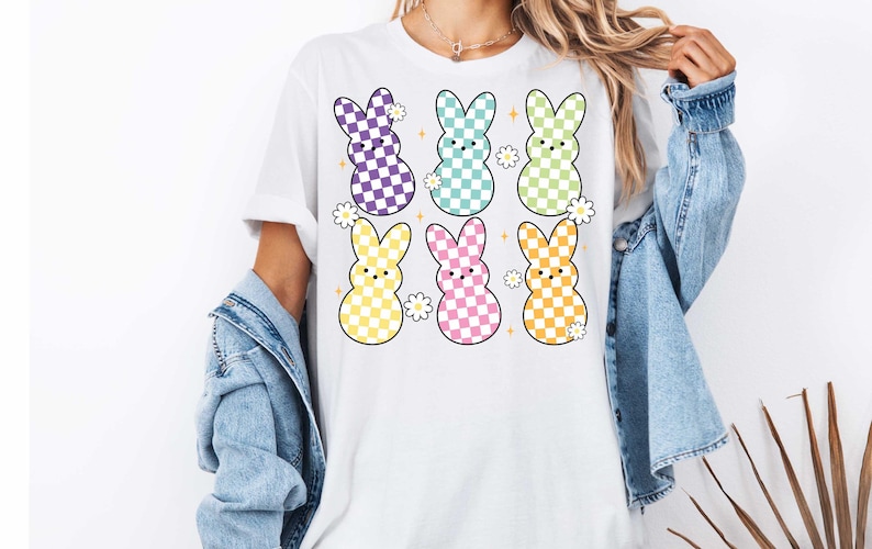 May include: White t-shirt with a graphic of six pastel colored Easter bunnies with checkered patterns. Each bunny has a small flower behind it.