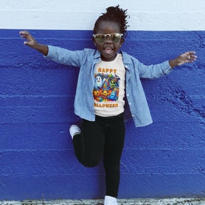 May include: A child wearing a white t-shirt with a colorful Halloween graphic and the text "Happy Halloween". The child is also wearing sunglasses, a denim jacket, and black pants.