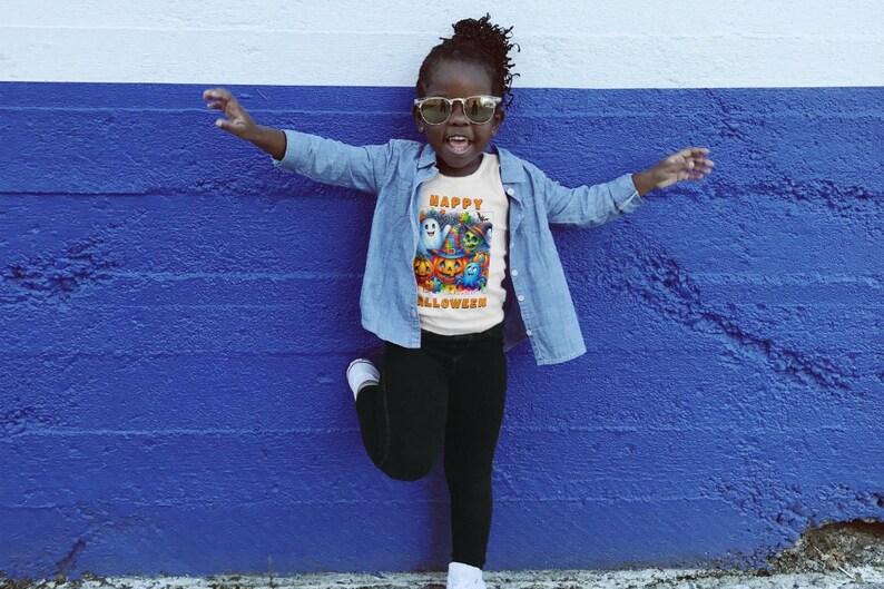 May include: A child wearing a white t-shirt with a colorful Halloween graphic and the text "Happy Halloween". The child is also wearing sunglasses, a denim jacket, and black pants.