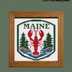 May include: A cross-stitch pattern in a wooden frame featuring a red lobster, green pine trees, and gray rocks above blue water, enclosed in a dark green border. The text MAINE is centered at the top, and PDF DOWNLOAD is printed above the frame.
