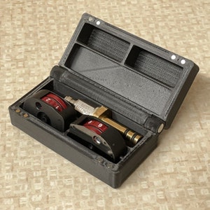 May include: A black plastic case with a hinged lid, containing two small, round, red and black numbered dials and a gold-coloured metal fitting labelled "BEST FITTINGS".