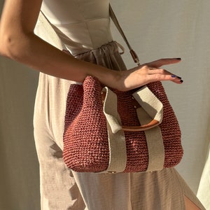 A dusty rose handmade crochet raffia purse bag with a woven-style crochet texture, beige fabric straps, brown leather accents, and a detachable shoulder strap, carried as a bohemian summer handbag.