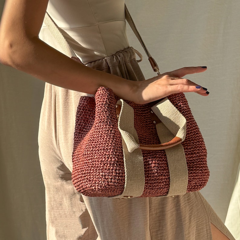 A dusty rose handmade crochet raffia purse bag with a woven-style crochet texture, beige fabric straps, brown leather accents, and a detachable shoulder strap, carried as a bohemian summer handbag.