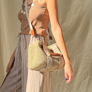 May include: A woman carrying a handmade crochet raffia purse bag with beige fabric straps, brown leather details, and a natural bohemian summer handbag style. The bag is worn as a crossbody purse with a lightweight casual look.