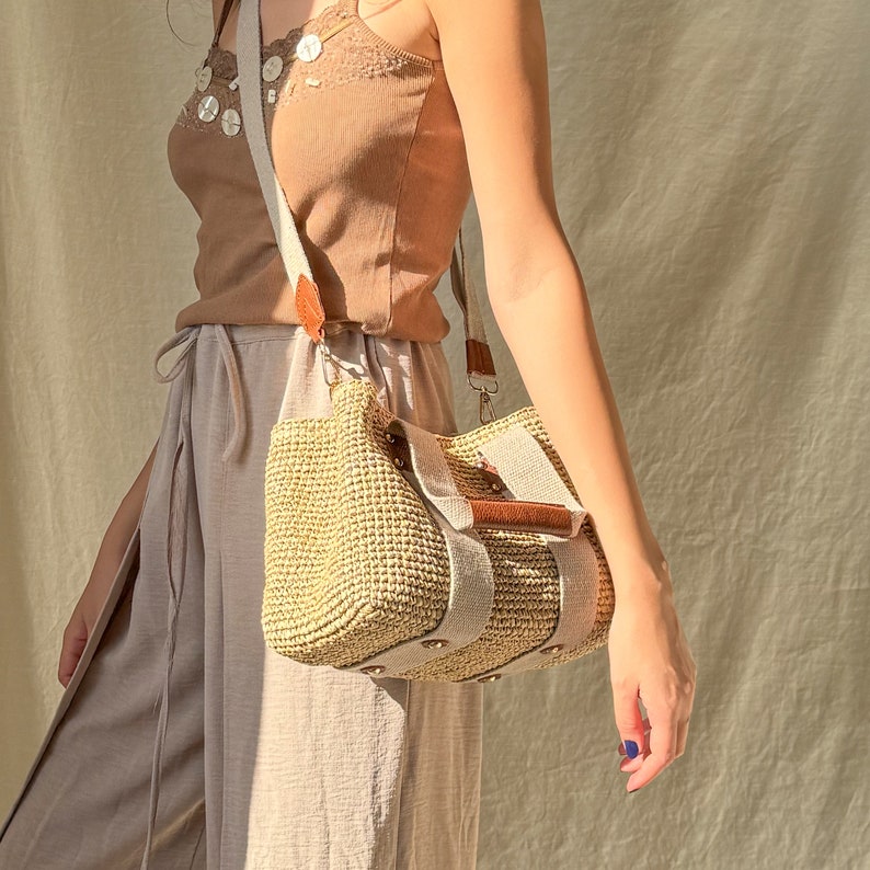 May include: A woman carrying a handmade crochet raffia purse bag with beige fabric straps, brown leather details, and a natural bohemian summer handbag style. The bag is worn as a crossbody purse with a lightweight casual look.