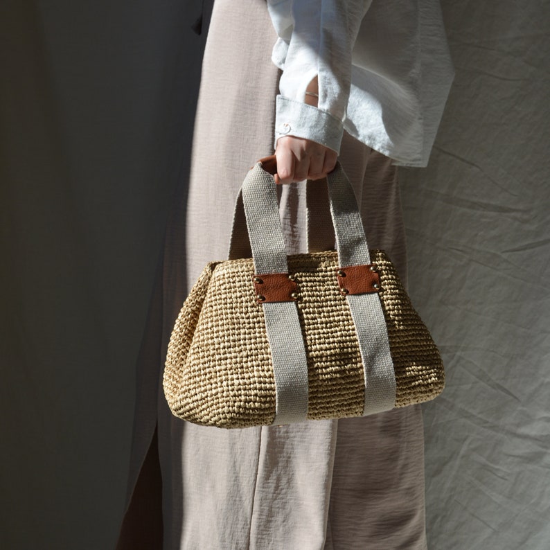 A beige handmade crochet raffia purse bag with a woven-style texture, brown leather accents, thick beige fabric handles, and a lightweight bohemian summer style for casual everyday outfits.