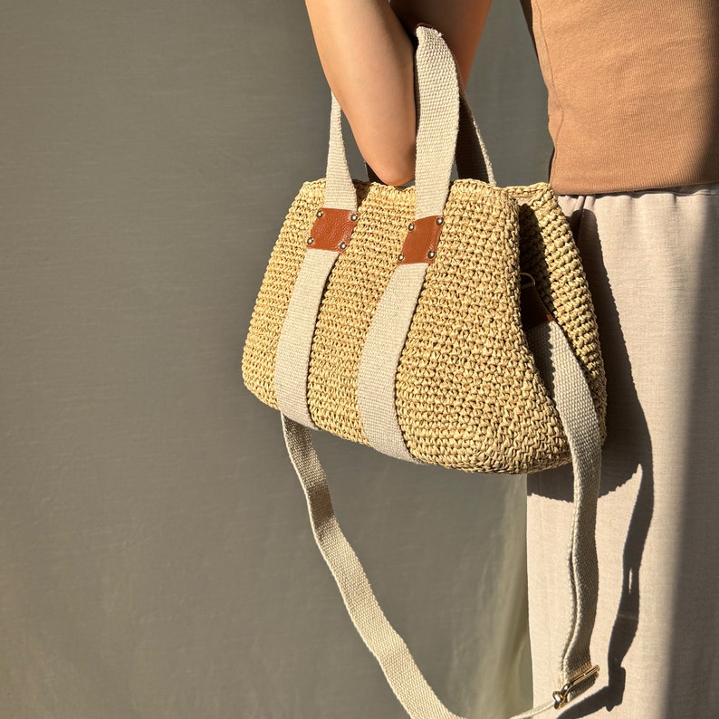 May include: A beige handmade crochet raffia purse bag with a woven-style crochet texture, brown leather accents, beige fabric handles, and a long adjustable strap.