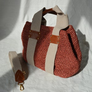 A dusty rose handmade crochet raffia purse bag with a woven-style crochet texture, cream fabric handles, brown leather accents, and a detachable shoulder strap.