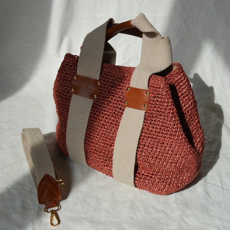 A dusty rose handmade crochet raffia purse bag with a woven-style crochet texture, cream fabric handles, brown leather accents, and a detachable shoulder strap.