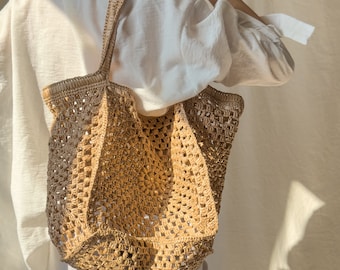 May include: A large, crocheted tote bag made of natural straw-like material. The bag has a square shape and a wide, open top. The bag is being held by a person wearing a white shirt.