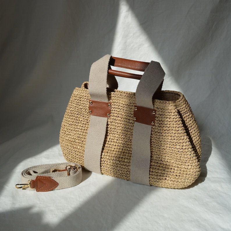 A beige handmade crochet raffia purse bag with a woven-style texture, brown leather handles, a detachable shoulder strap, and gold-tone hardware accents for a natural bohemian summer handbag style.