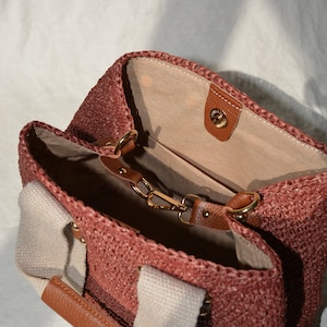 May include: A dusty rose handmade crochet raffia purse bag with beige canvas lining, brown leather details, an inner pocket, and a natural bohemian summer handbag style.