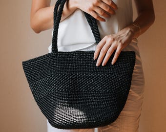 May include: A black hand-crocheted tote bag featuring a structured, wide-base design with two integrated top handles. The bag is crafted from a textured, woven material with a consistent dark finish and a subtle horizontal pattern across the body.