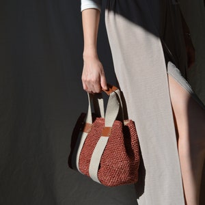 A red handmade crochet raffia purse bag with a woven-style texture, brown leather accents, and a natural bohemian summer style, held by a person wearing a light-colored dress.