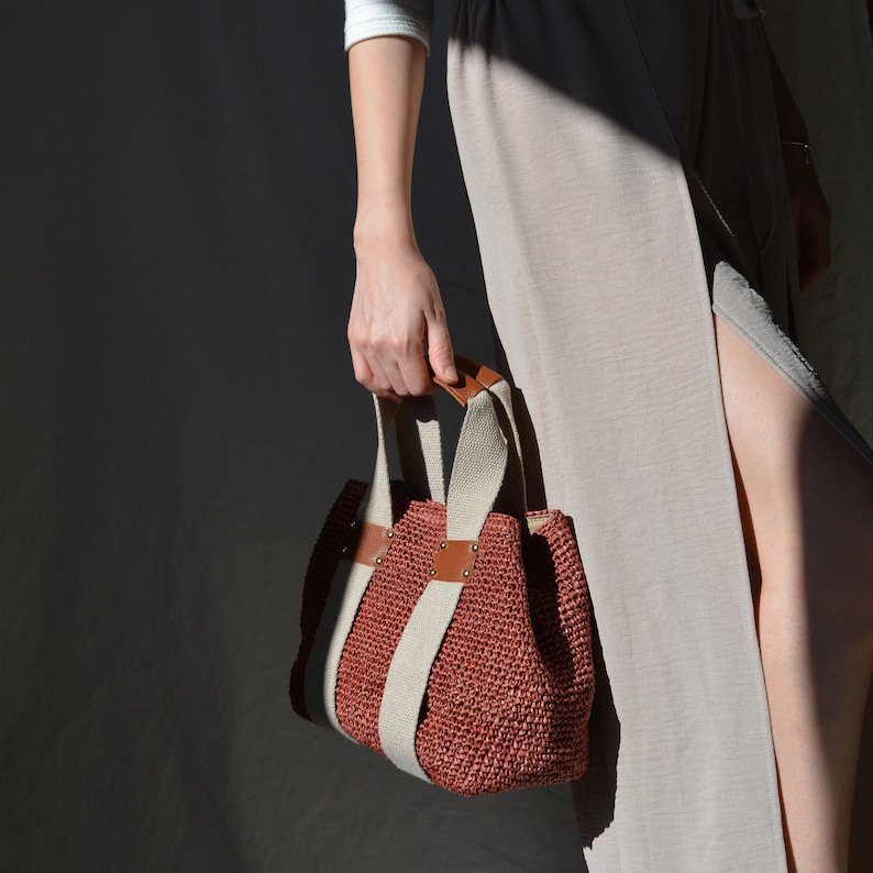 A red handmade crochet raffia purse bag with a woven-style texture, brown leather accents, and a natural bohemian summer style, held by a person wearing a light-colored dress.