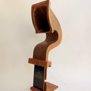 May include: A wooden phone stand with a unique, curved design. The stand is made of light brown wood and features a rectangular base, a curved support, and a top section designed to hold a phone. The stand is designed to hold a phone vertically.