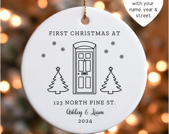 May include: A white ceramic Christmas ornament with a gold string. The ornament features a black line drawing of a door, two Christmas trees, snowflakes, and the text "FIRST CHRISTMAS AT", "123 NORTH PINE ST.", "Ashley & Liam", and "2024".