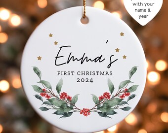 May include: A white ceramic Christmas ornament with the text "Emma's First Christmas 2024". The ornament is decorated with green leaves, red berries, and gold stars. A gold string is attached for hanging.