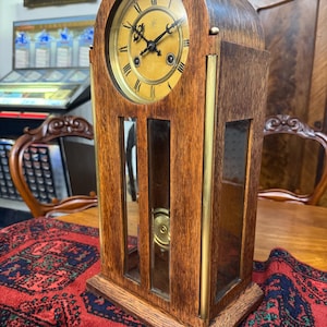 Peut inclure: Une horloge en bois vintage avec un cadran rond et des chiffres romains. L'horloge a un cadran brun clair avec des aiguilles noires et un cadre doré. Le boîtier en bois de l'horloge est de couleur marron foncé avec des accents en laiton et des panneaux de verre.