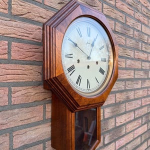 Puede incluir: Un reloj de pared de madera con una esfera clara y números romanos negros. El reloj tiene un marco octogonal marrón oscuro y una ventana de péndulo cubierta de vidrio. El reloj está montado en una pared de ladrillo rojo.