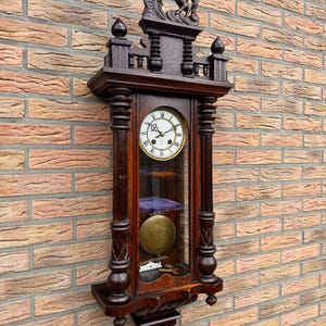 May include: A dark wood wall clock featuring a horse carving. The clock has a round face with Roman numerals, a pendulum, and a glass front. The clock is mounted on a brick wall. The clock's dimensions are approximately 76 cm in height.