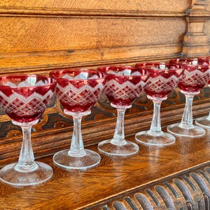Op de afbeelding: Een set van zes robijnrode kristallen likeurglazen met heldere stelen en voetjes. Elk glas heeft een gesneden, geometrisch patroon rond de kom. De glazen zijn gerangschikt op een gepolijst houten oppervlak, wat hun elegante ontwerp benadrukt.