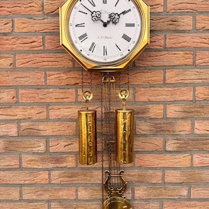 Puede incluir: Un reloj de pared dorado con esfera blanca y números romanos negros. El reloj tiene un péndulo y dos pesas cilíndricas. La esfera del reloj dice "Villain" y "A. St. Reitz". El reloj está colgado en una pared de ladrillo.