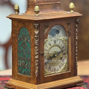May include: Antique wooden clock with a brass face and ornate carvings. The clock has a glass front and a green and gold design on the side.