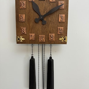 Puede incluir: Un reloj de madera con marcadores de hora en números romanos y manecillas negras. El reloj tiene un acabado de madera oscura y detalles en latón. Dos pesas negras cuelgan de cadenas.