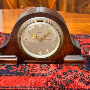 May include: A vintage wooden clock with a round face and gold-colored hands and numerals. The clock has a dark brown, arched wooden frame and sits on a red and blue patterned rug. The clock face has the word "WUBA" printed on it.