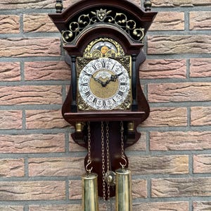 Op de afbeelding: Een wandklok van donker hout met een witte wijzerplaat met zwarte Romeinse cijfers, een goudkleurig midden en een maanfase-indicator. De klok heeft twee koperen gewichten aan kettingen, decoratieve koperen ornamenten en metalen versieringen.