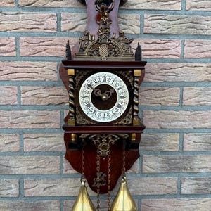 May include: A dark wood wall clock with a white face and Roman numerals. The clock has gold-colored weights and a decorative top with a figure holding a globe. The clock is mounted on a brick wall.
