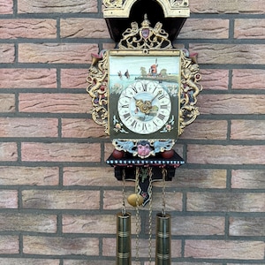 Puede incluir: Un reloj de pared decorativo con un marco dorado detallado y una escena pintada en la esfera. El reloj tiene un péndulo y pesas, y está montado en una pared de ladrillo.